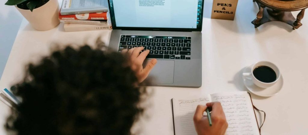 Author writing in a notebook beside a laptop and coffee