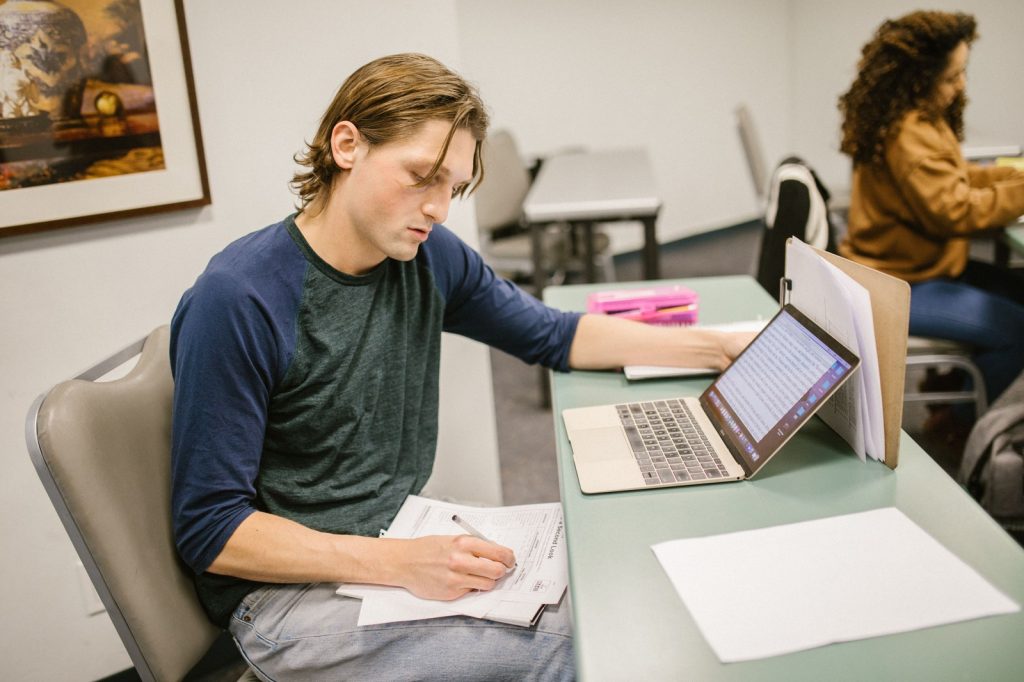 Student writing an essay at a desk with a laptop in a classroom