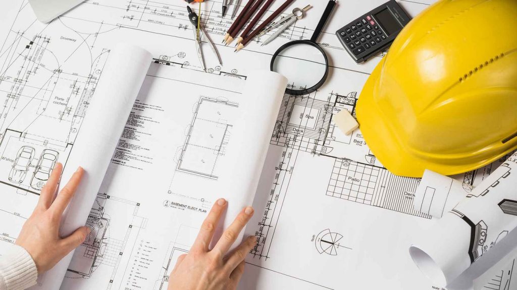 Hands reviewing construction blueprints with a hard hat, calculator and measuring tools for a takeoff