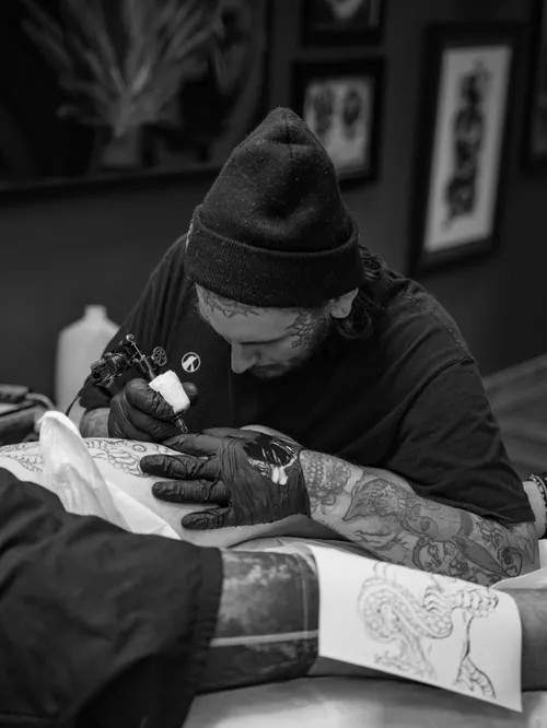 Tattoo artist in a beanie and gloves tattooing a client's arm in a studio